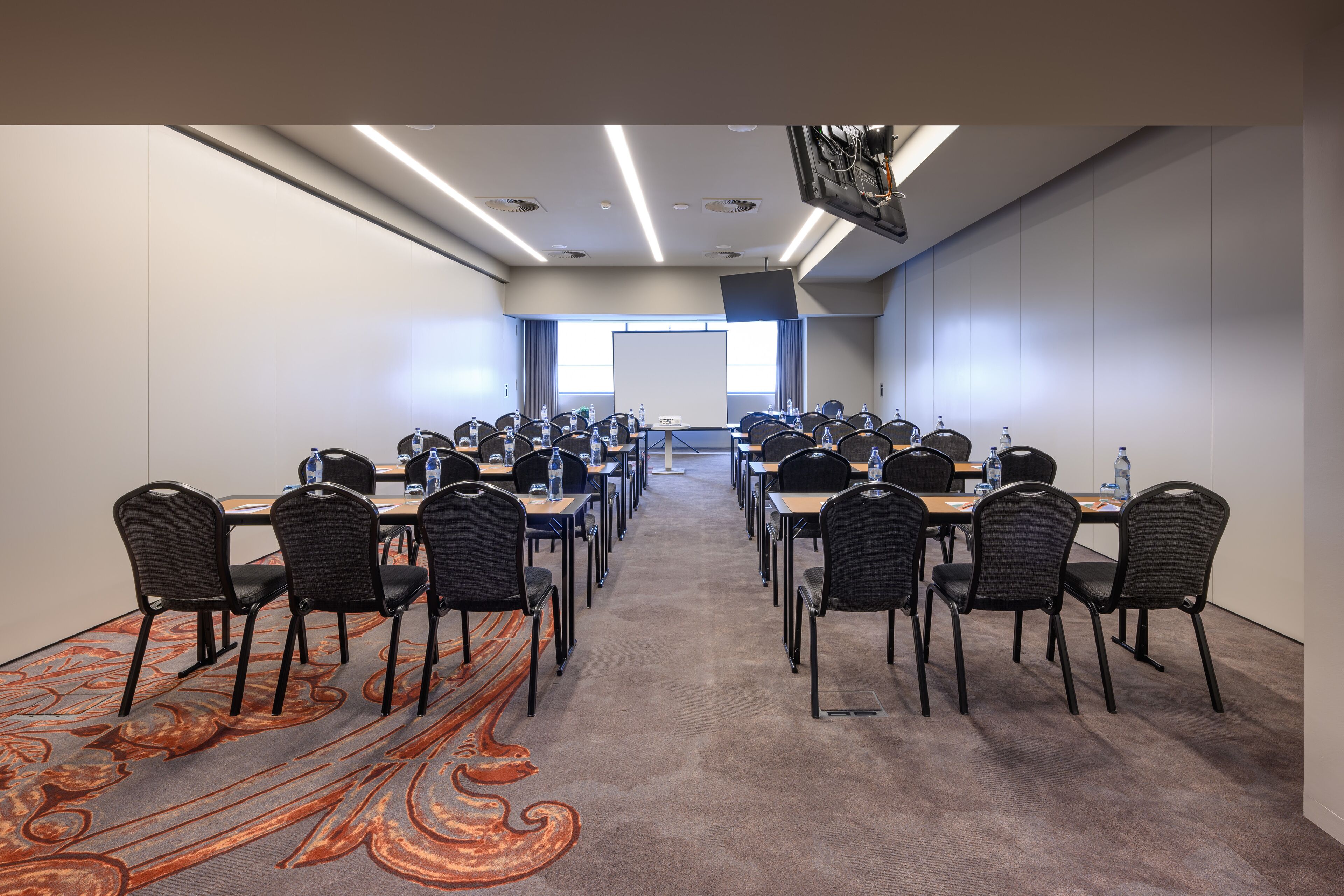 Un salón de conferencias con filas de mesas y sillas, una pantalla de proyector y suelo alfombrado.
