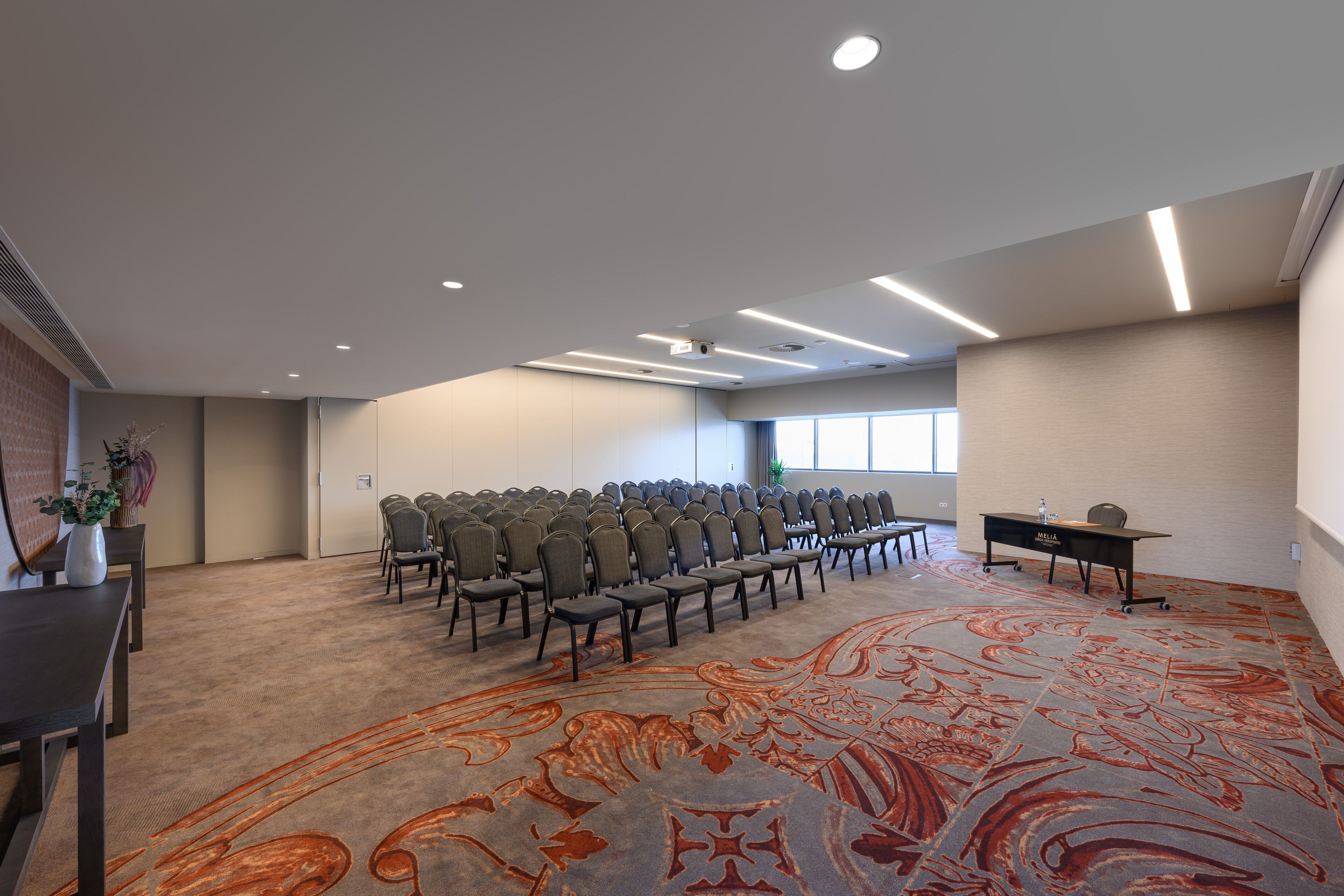 Un salón de conferencias con filas de sillas, piso alfombrado y un escritorio con una planta.