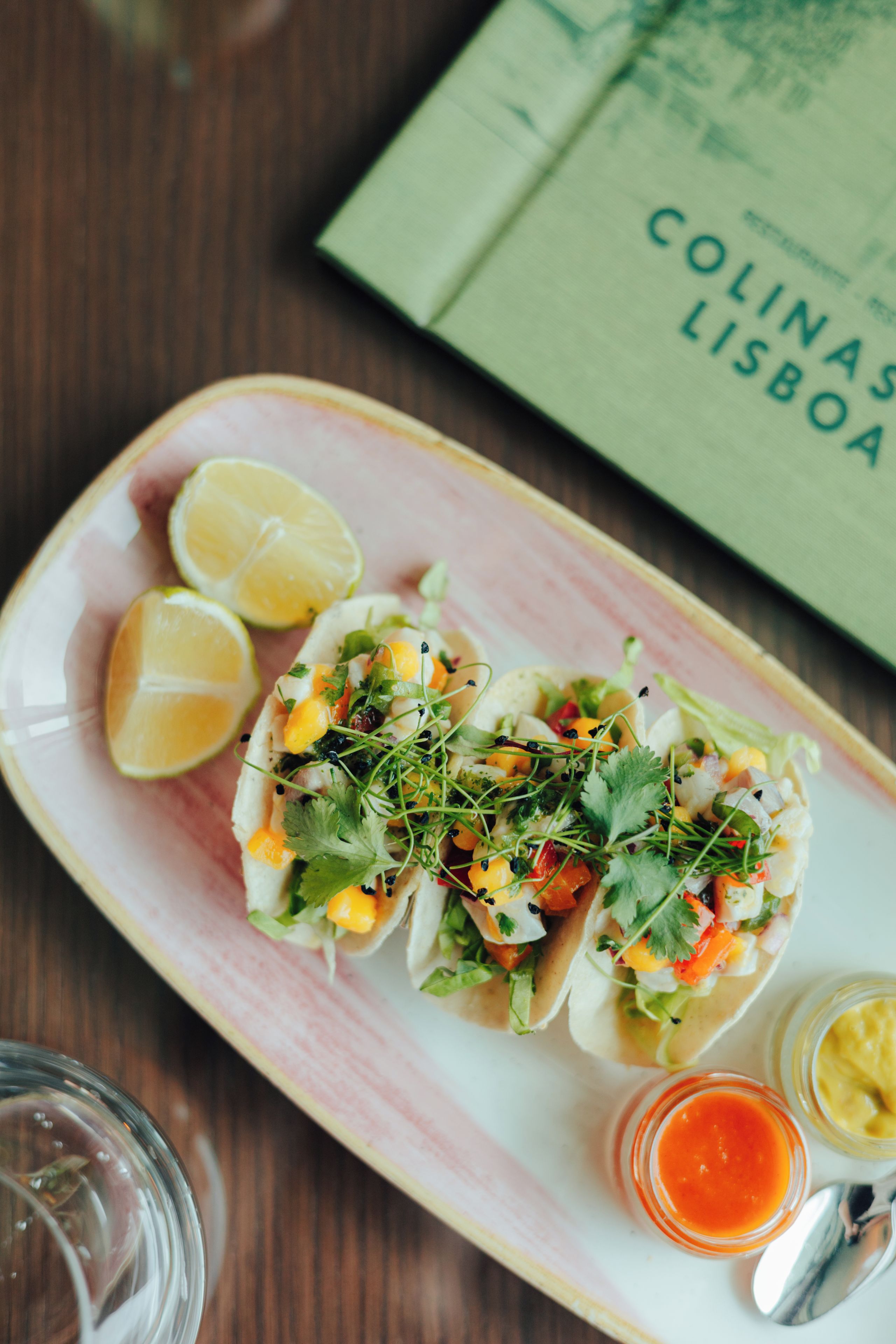 Un plato con dos tacos, un vaso de agua y un libro titulado 'Colinas Lisboa' sobre una mesa de madera.