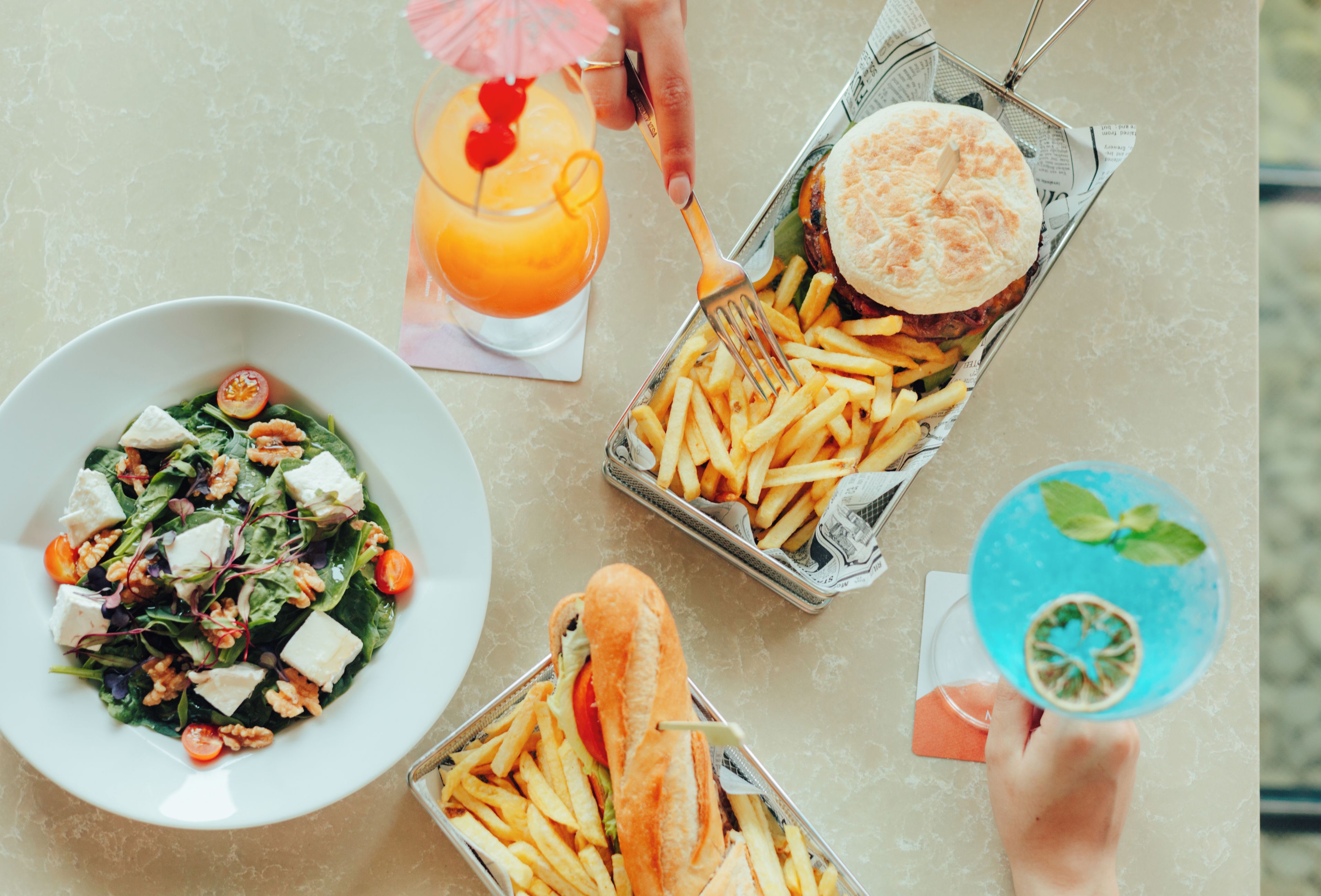 Una mesa con ensalada, hamburguesa, papas fritas, una bebida y postre.