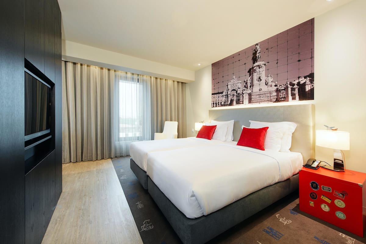 Ein Hotelzimmer mit einem großen Bett, einem Schreibtisch mit Lampe, einem Fernseher und einer Wand mit Stadtbildgemälde.