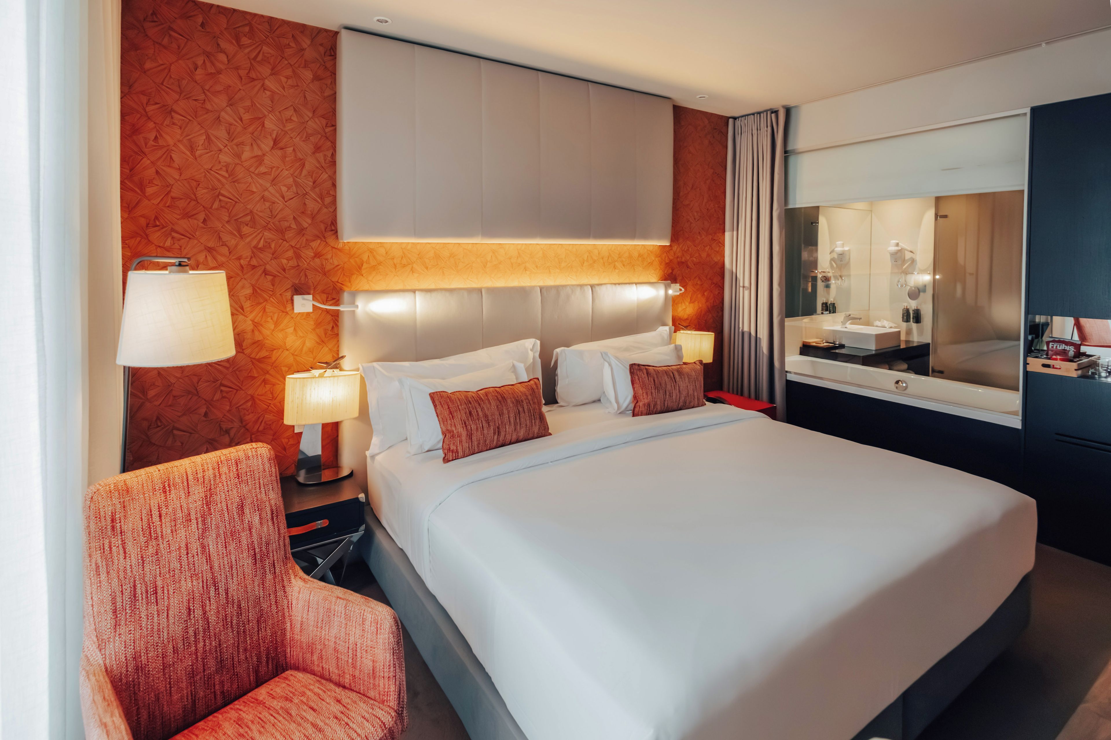 A hotel room with a large bed, two bedside lamps, a nightstand, a chair, a mirror, and a window with curtains.