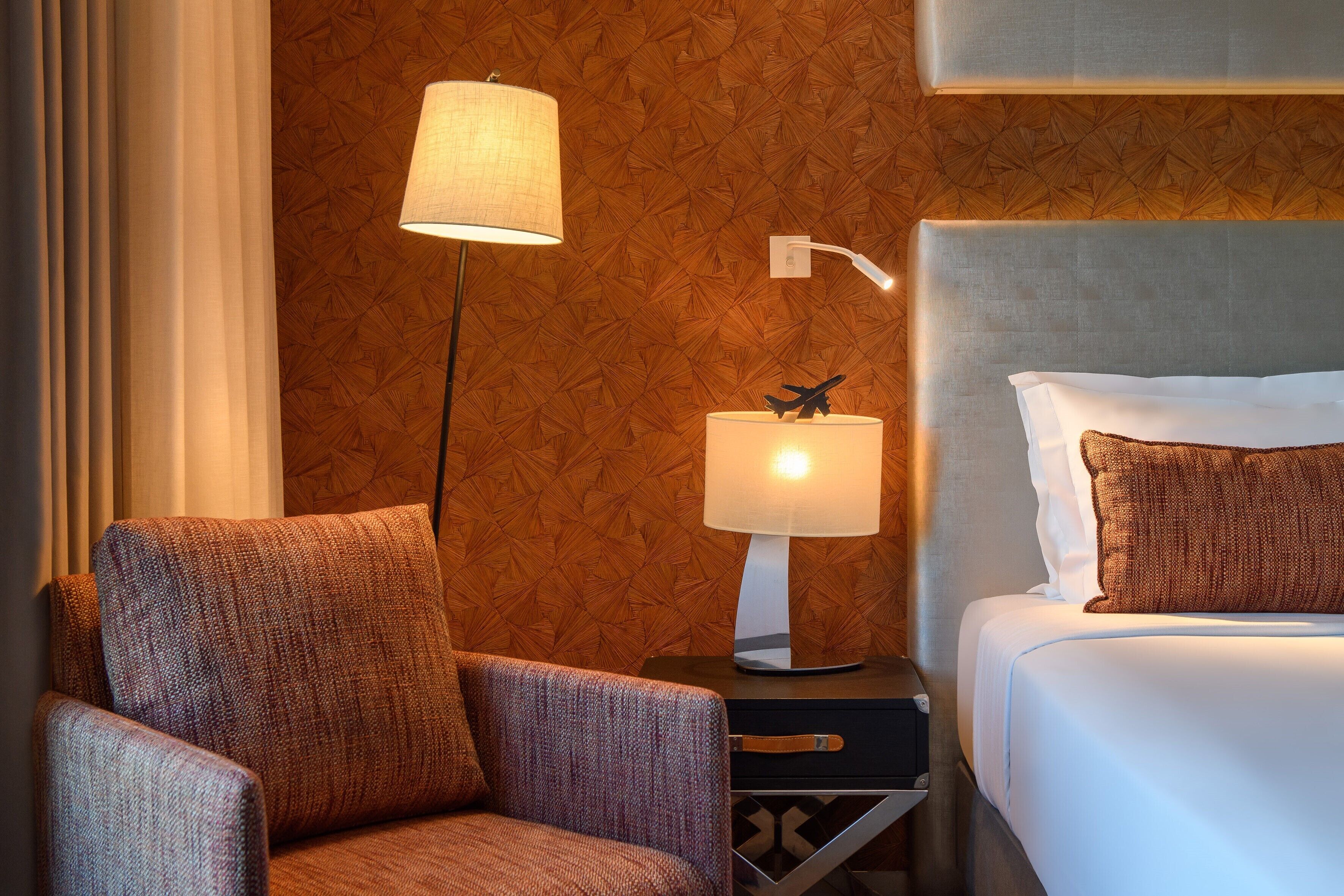 Quarto de hotel com uma cama, uma mesa de cabeceira com luminária, uma cadeira e parede com padrão geométrico.