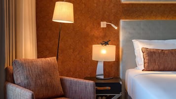 Quarto de hotel com uma cama, uma mesa de cabeceira com luminária, uma cadeira e parede com padrão geométrico.
