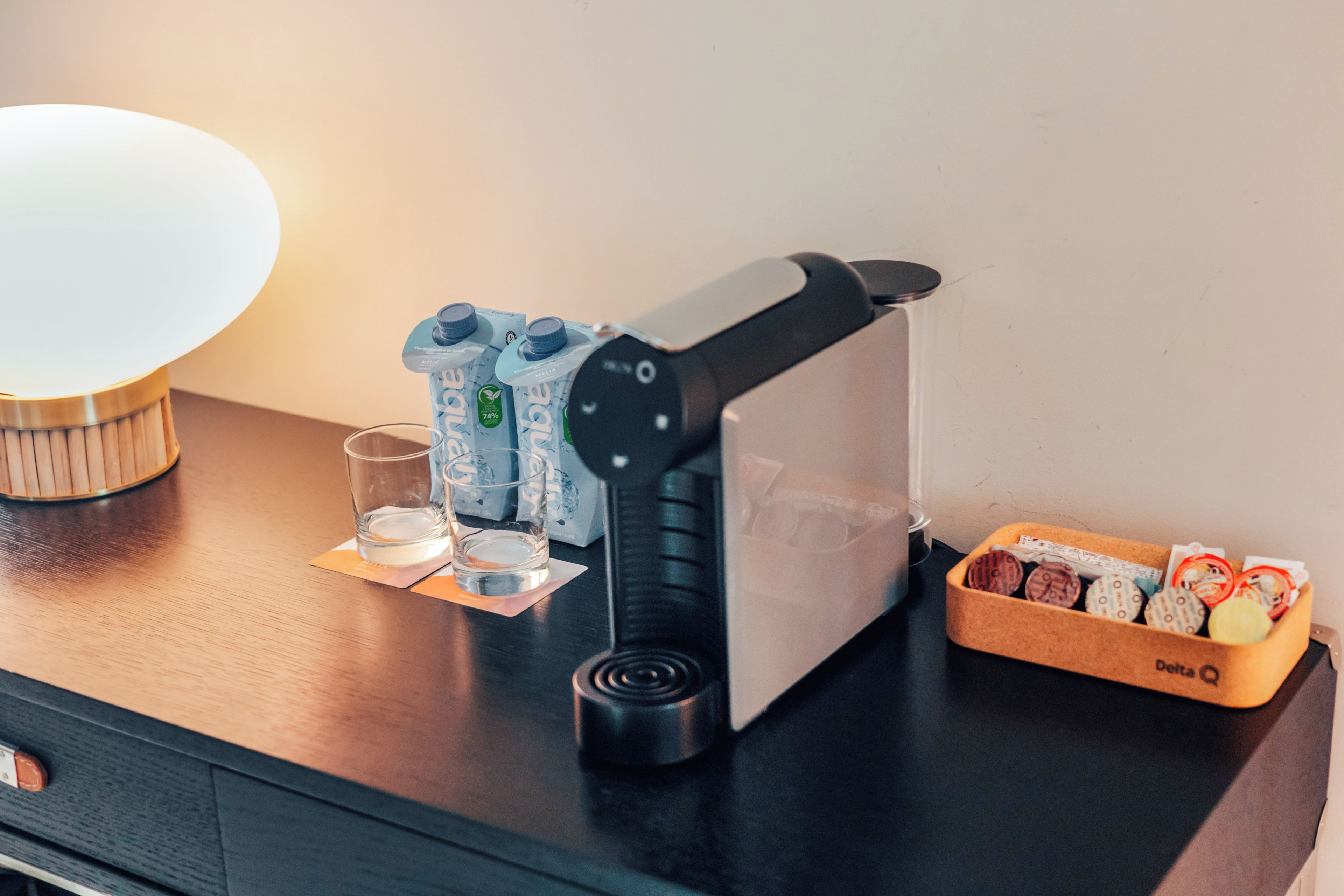 A coffee machine on a wooden table with a glass, a pack of Moka pots, and a Delta Q container with coffee capsules.