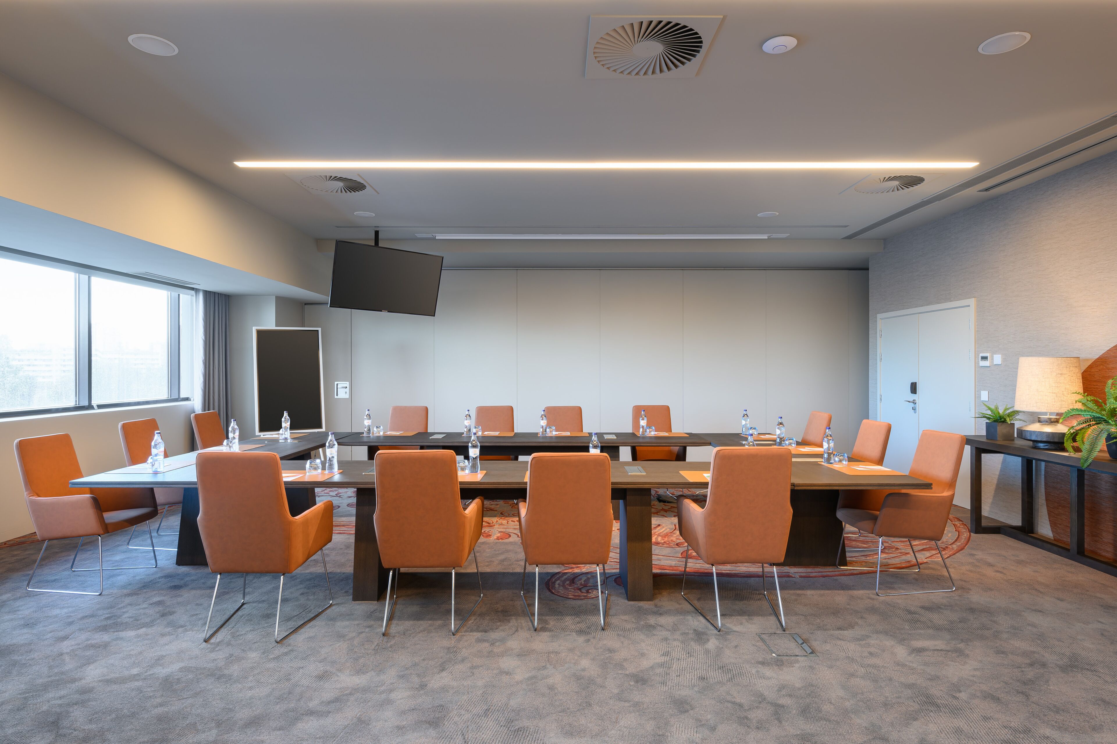Een vergaderzaal met een lange tafel, oranje stoelen en een flatscreen televisie.