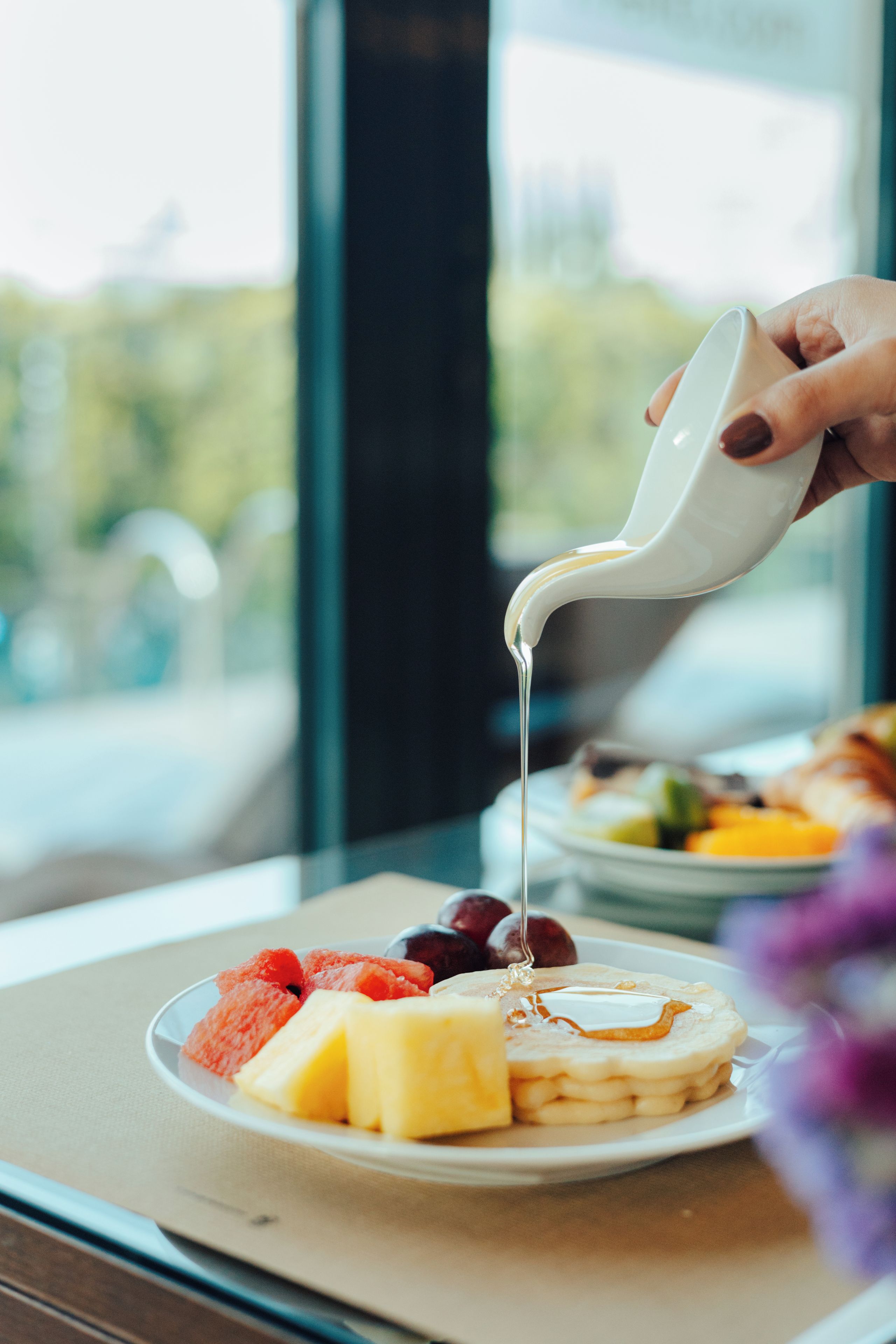 A hand pouring syrup over a plate of pancakes with fruit and a glass of milk.