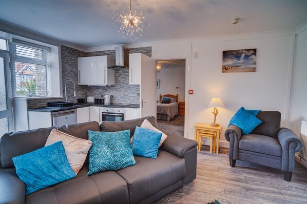 A modern living room with a sofa and armchair, a kitchen in the background, and a bedroom visible through an open door. - Beachmount Holiday Apartments (Colwyn Bay)