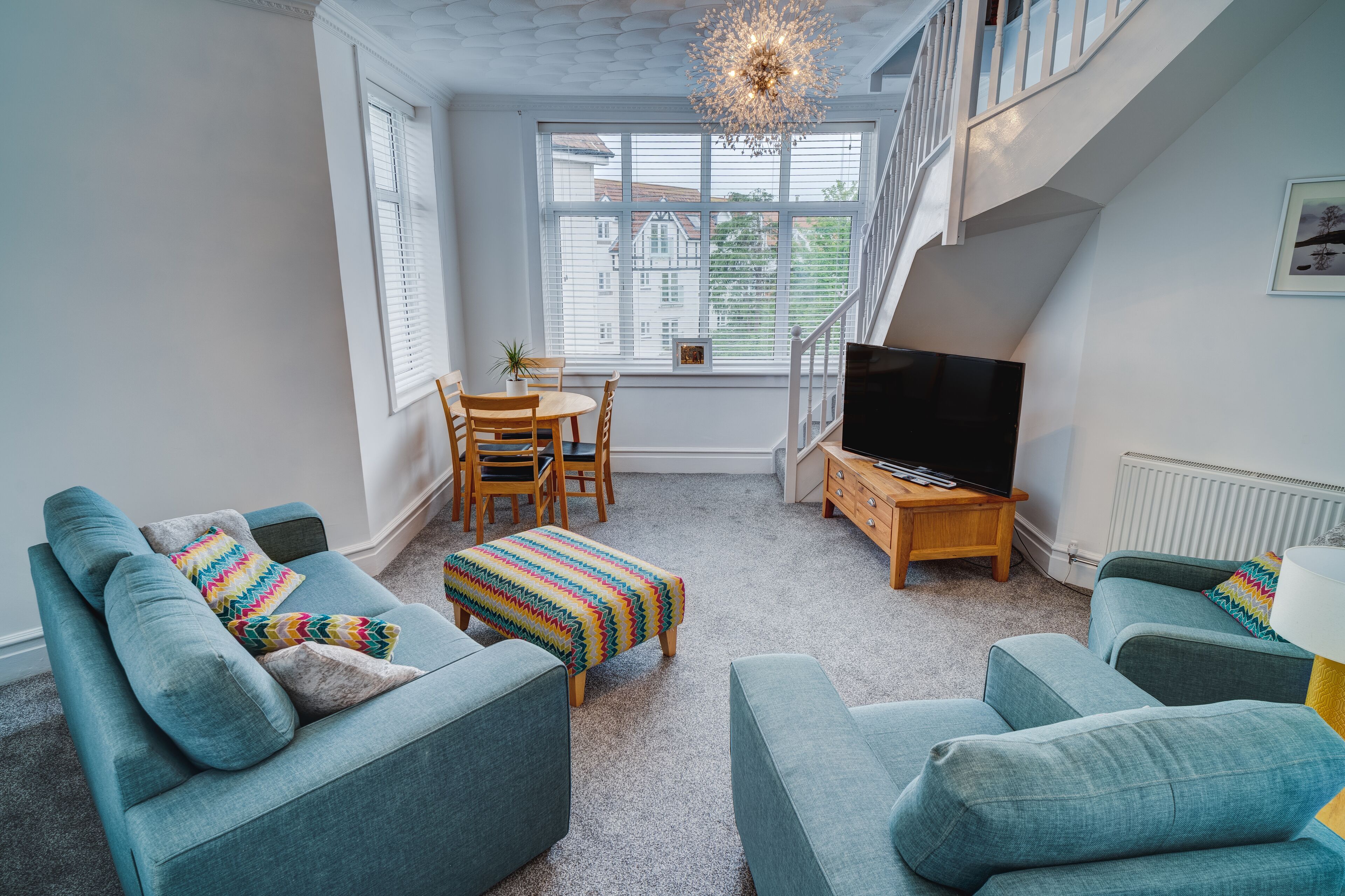 A living room with teal sofas, a wooden coffee table, and a TV. There is a staircase with a railing leading to an upper level.