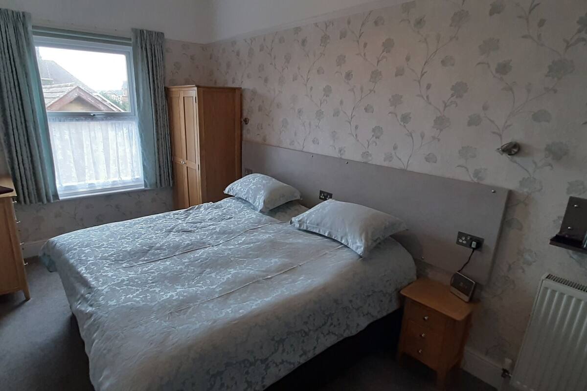 Une chambre à coucher comprenant un lit, une armoire, un téléviseur fixé au mur, une fenêtre avec des rideaux et un radiateur.