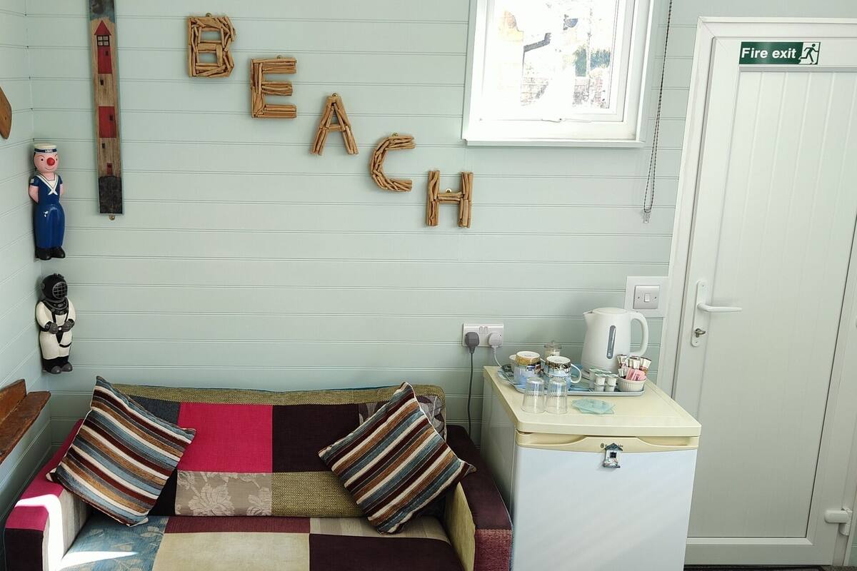 Une pièce chaleureuse avec un canapé, des coussins, une petite table avec une bouilloire et un mur sur lequel est écrit le mot « PLAGE ».