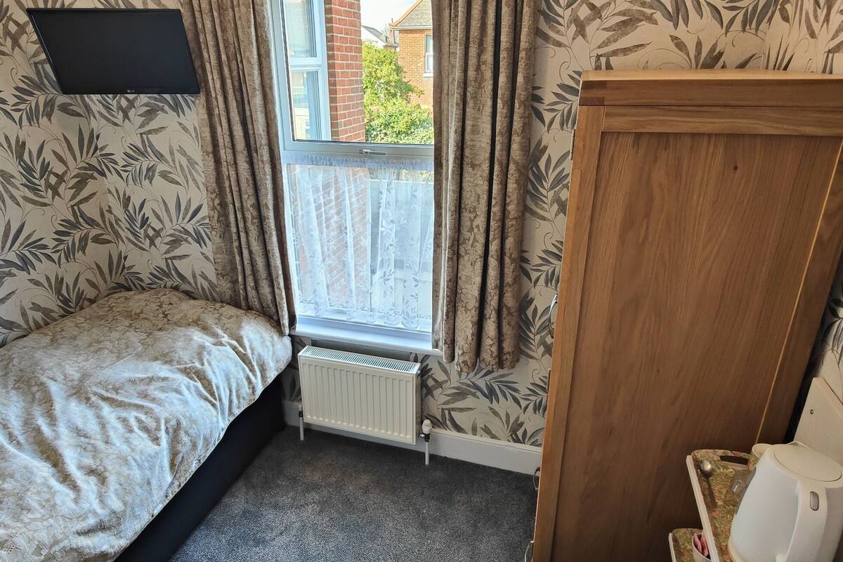 Une petite chambre avec un lit simple, un radiateur, une armoire en bois et une fenêtre avec des rideaux.