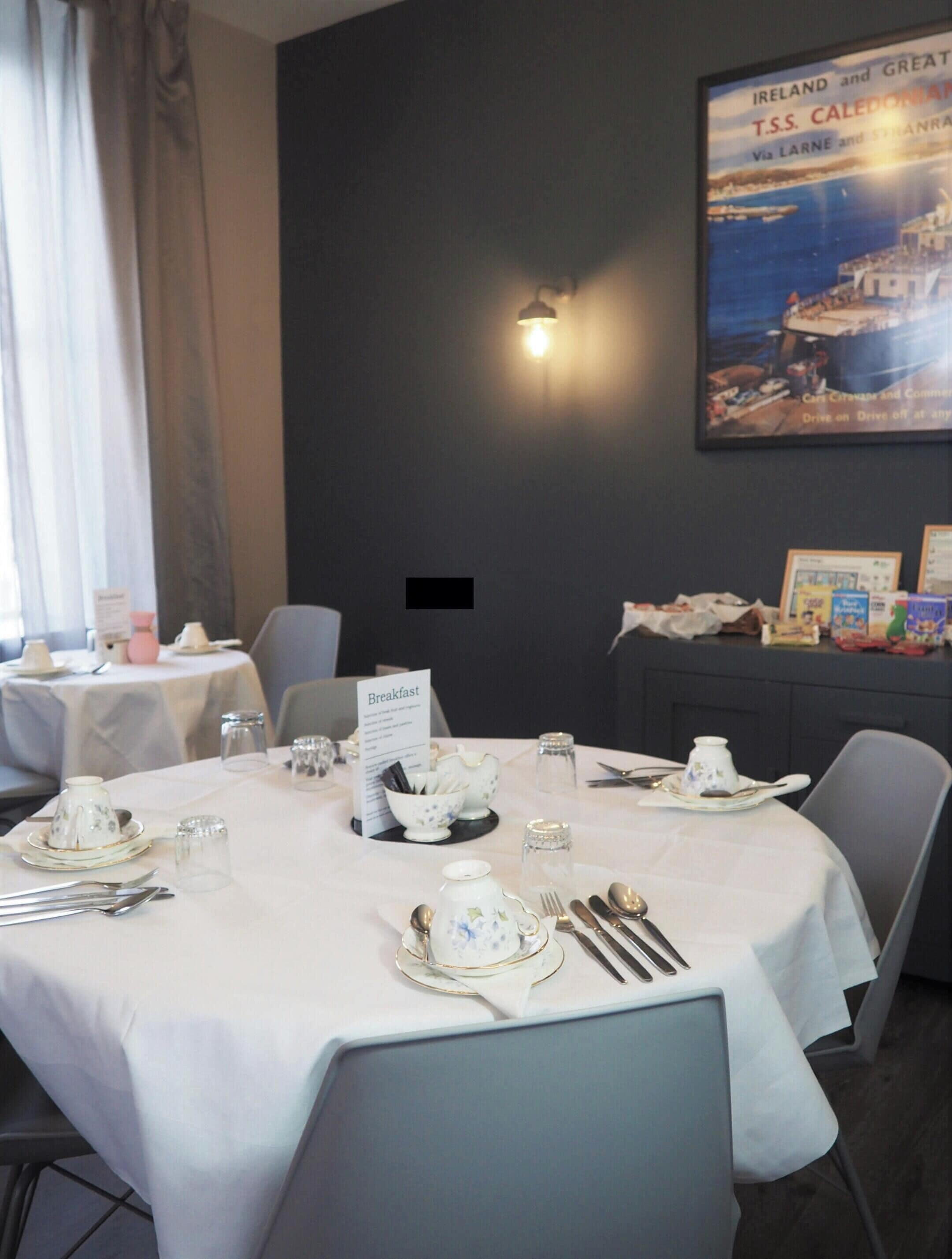 A dining area with round tables set for breakfast, featuring white tablecloths, floral-patterned tea sets, and silverware.