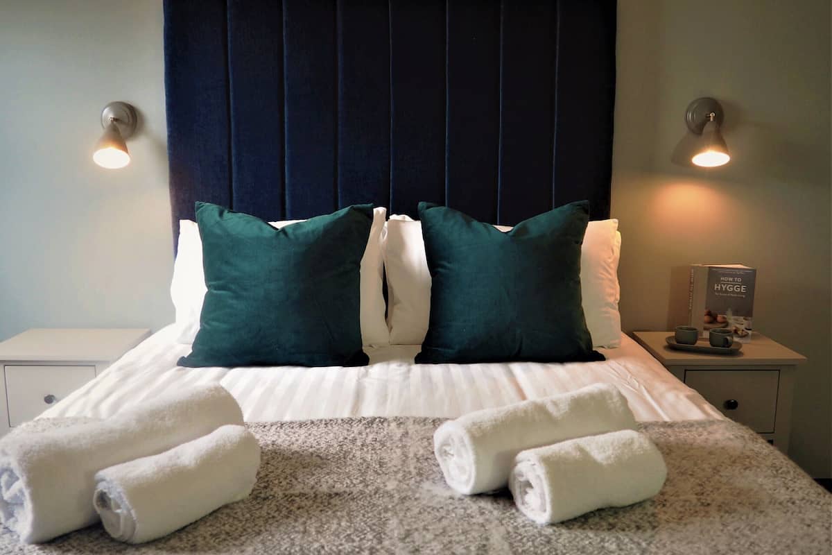 A neatly made bed with white linens, two green and one white pillow, and rolled white towels on a textured grey carpet.