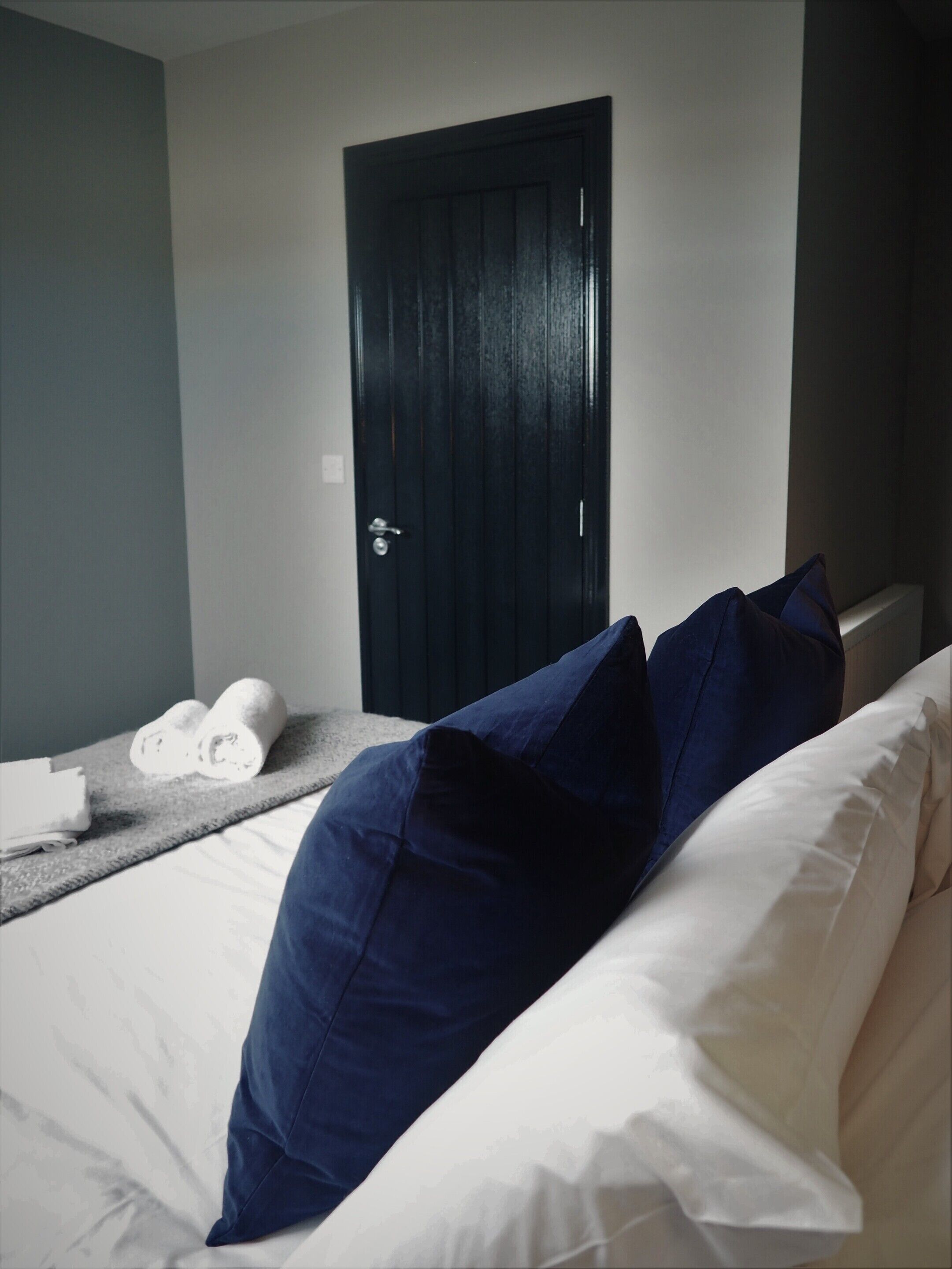 A hotel room with a bed, a dark door, and a towel rack with towels.