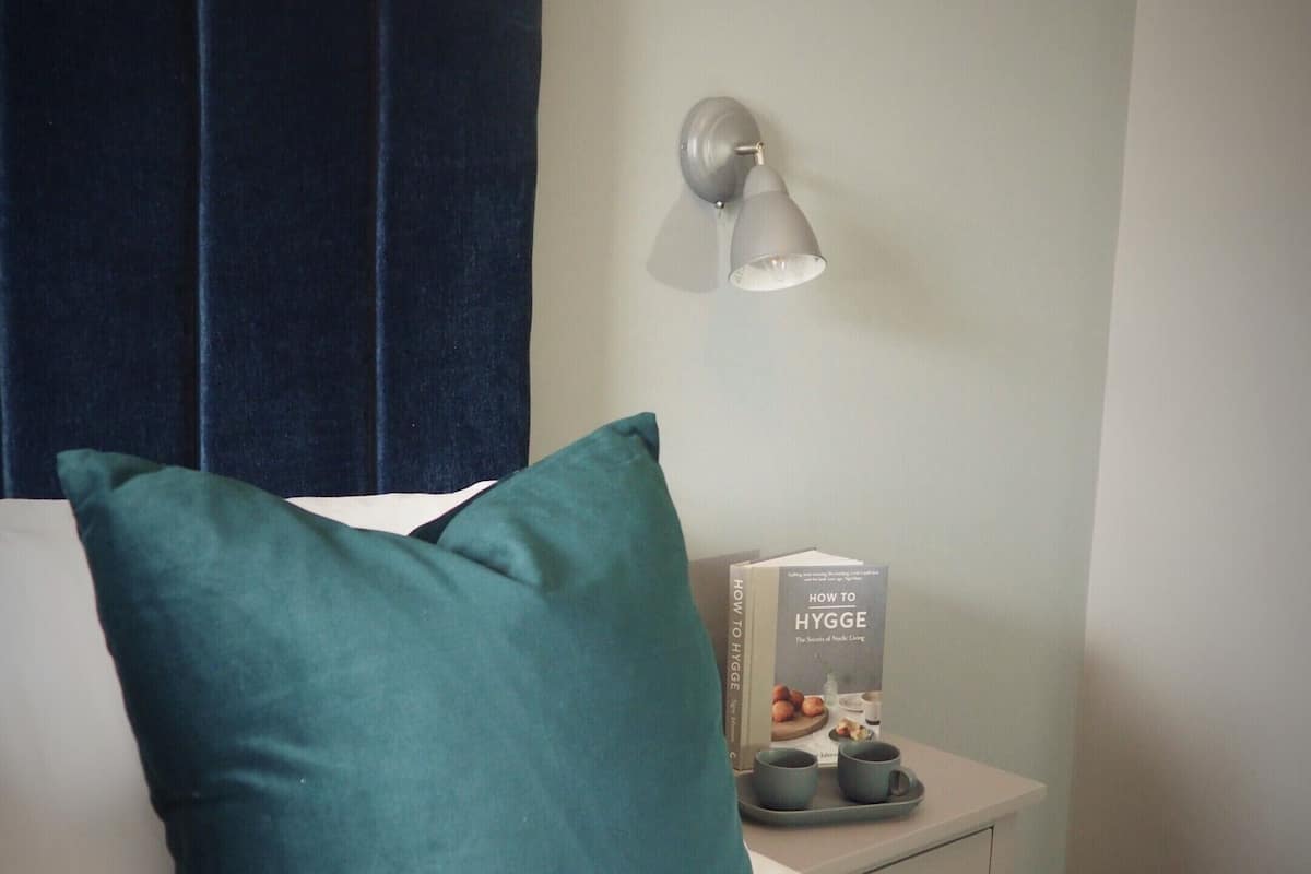 A hotel room with a bed, a teal pillow, a bedside table with a book, a cup, and a tray.