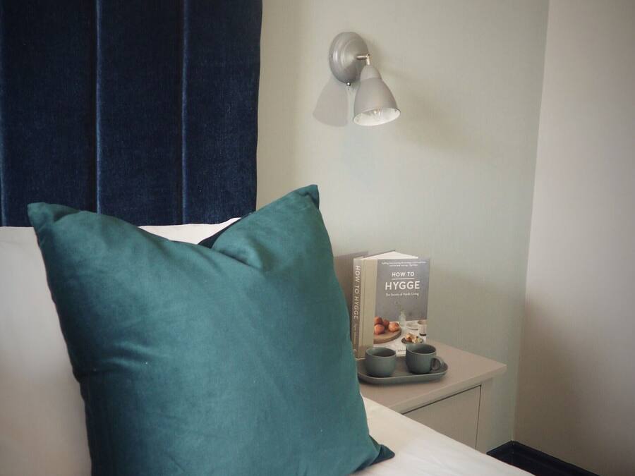A hotel room with a bed, a teal pillow, a bedside table with a book, a cup, and a tray.