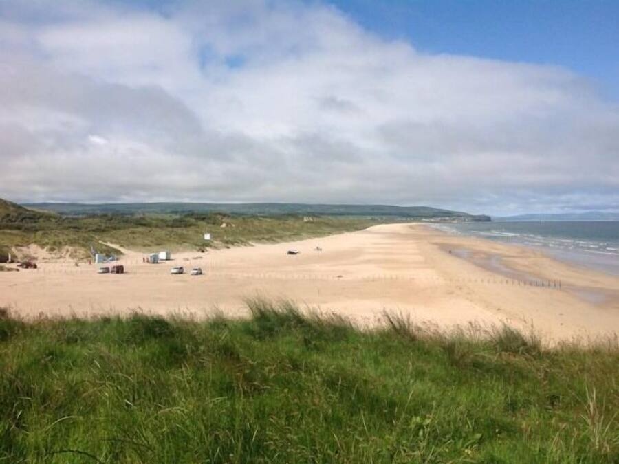 A beach with a sandy shore, grassy dunes, and a few structures in the distance.