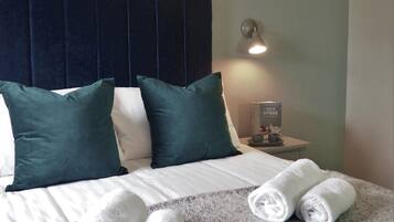 Una cama bien hecha con sábanas blancas y dos toallas enrolladas, un cabecero oscuro, y una mesita de noche con una lámpara y un libro titulado 'HOW TO HYGGE'.