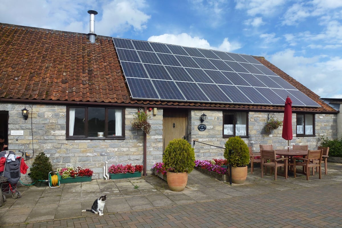 A stone building with a tiled roof, solar panels, a wooden door, and a brick patio with outdoor furniture and potted plants.