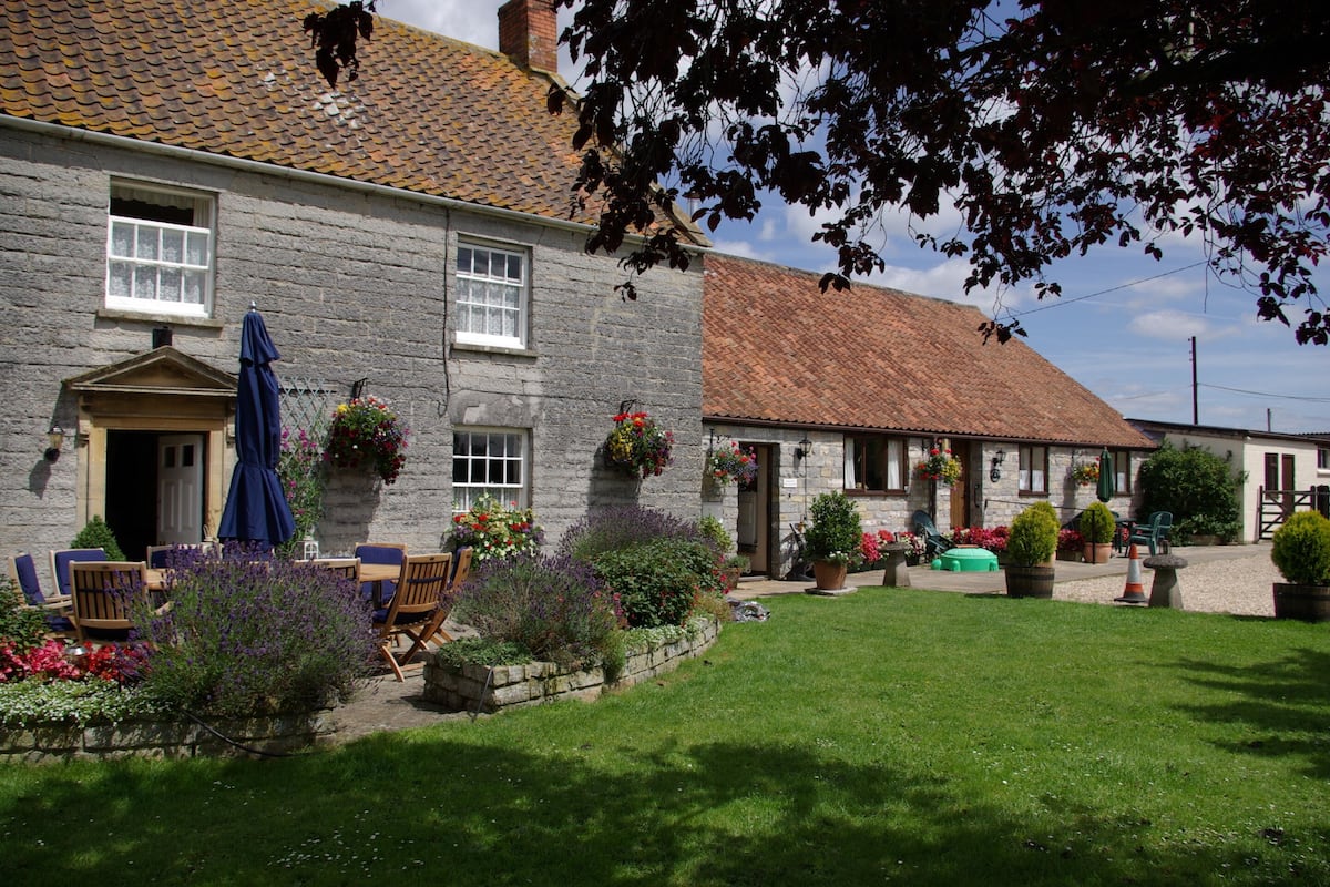 A stone building with a tiled roof, a front door, and a small garden with chairs and tables.