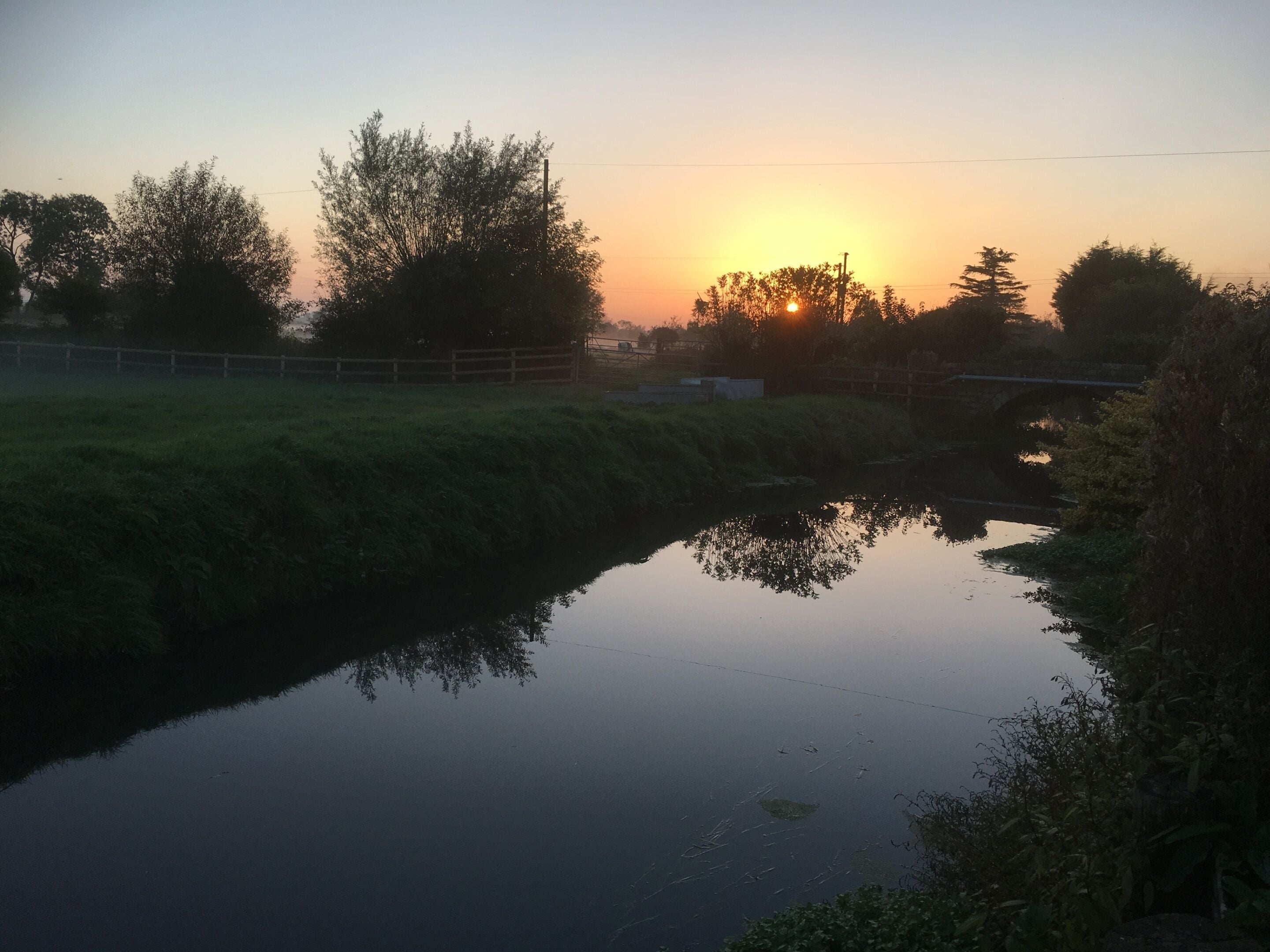 A calm river reflecting the sunset, a bridge, and surrounding greenery.
