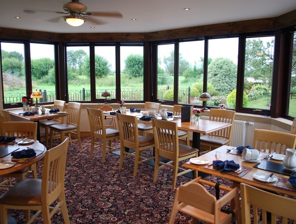 A dining area with wooden tables and chairs, large windows with a view of greenery, and a ceiling fan.
