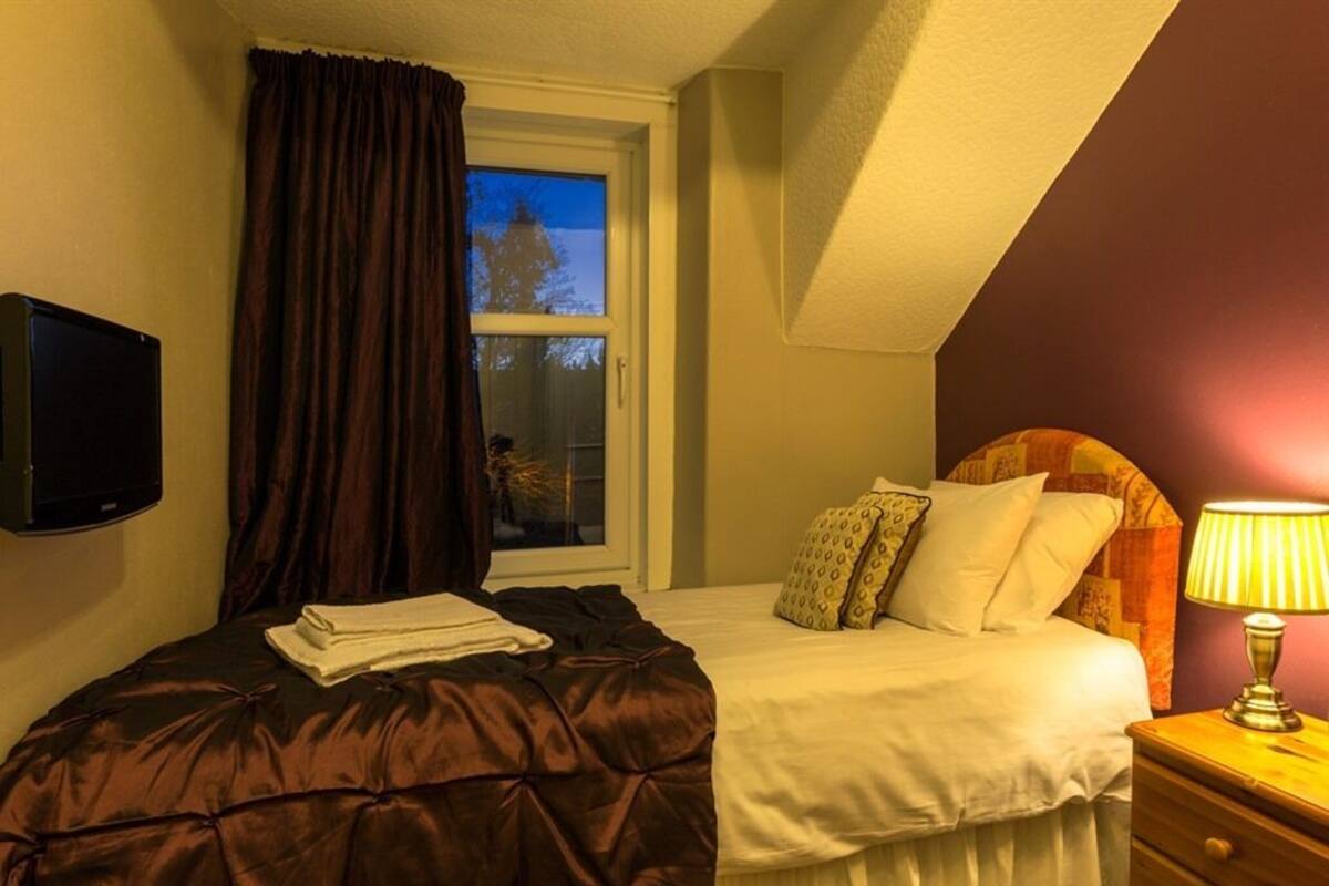 A bedroom with a bed, a window with curtains, a bedside lamp, and a TV on the wall.