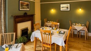 A dining area with wooden chairs and tables, a fireplace, and framed pictures on the walls. - Craglands Guest House (Keswick)