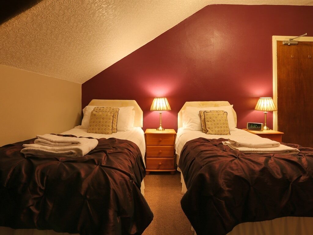 Habitación con dos camas, cada una con una mesita de noche y una lámpara. La habitación tiene techo inclinado y una puerta a la derecha.