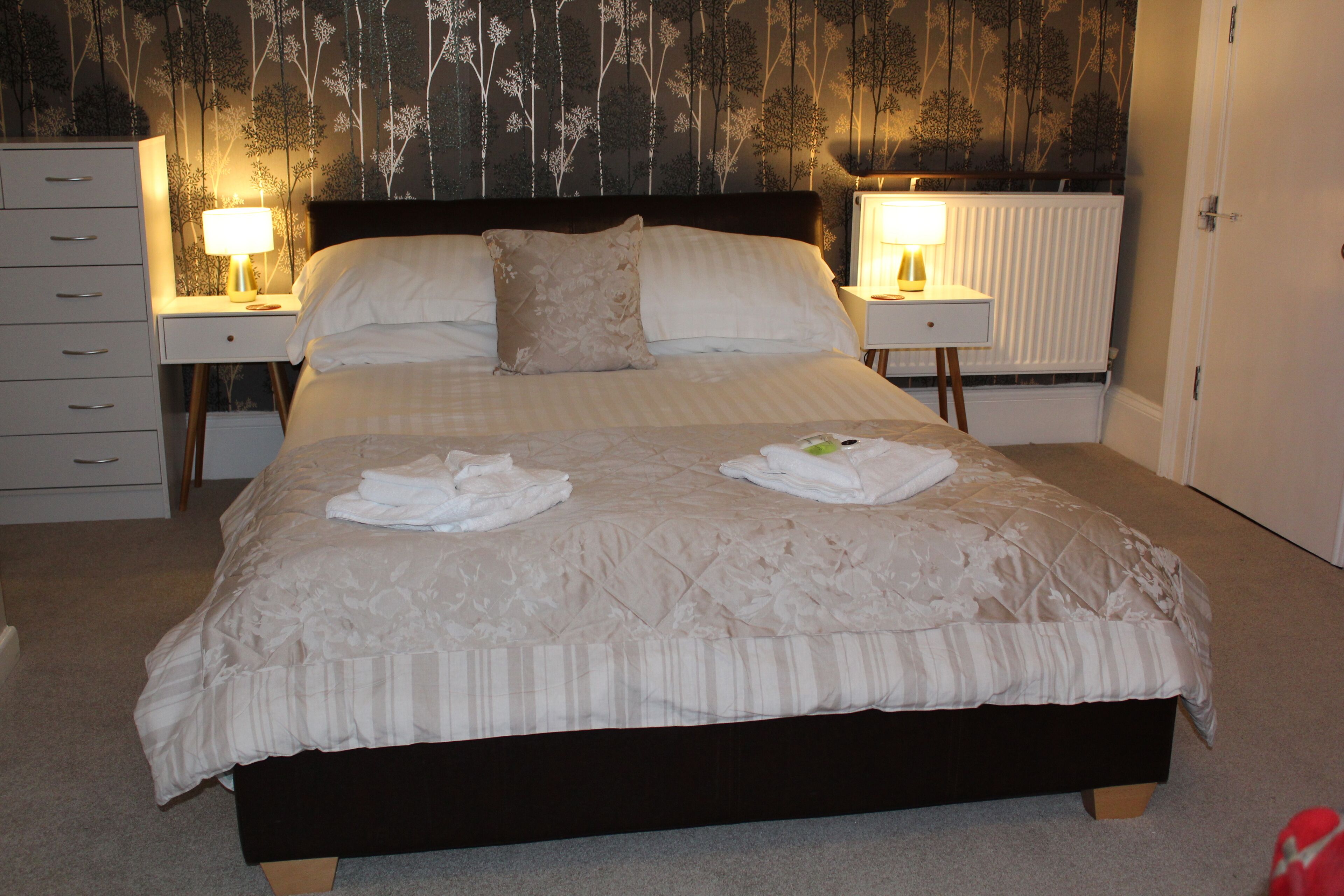 A neatly made bed with a dark headboard, two bedside lamps, and a radiator behind it.