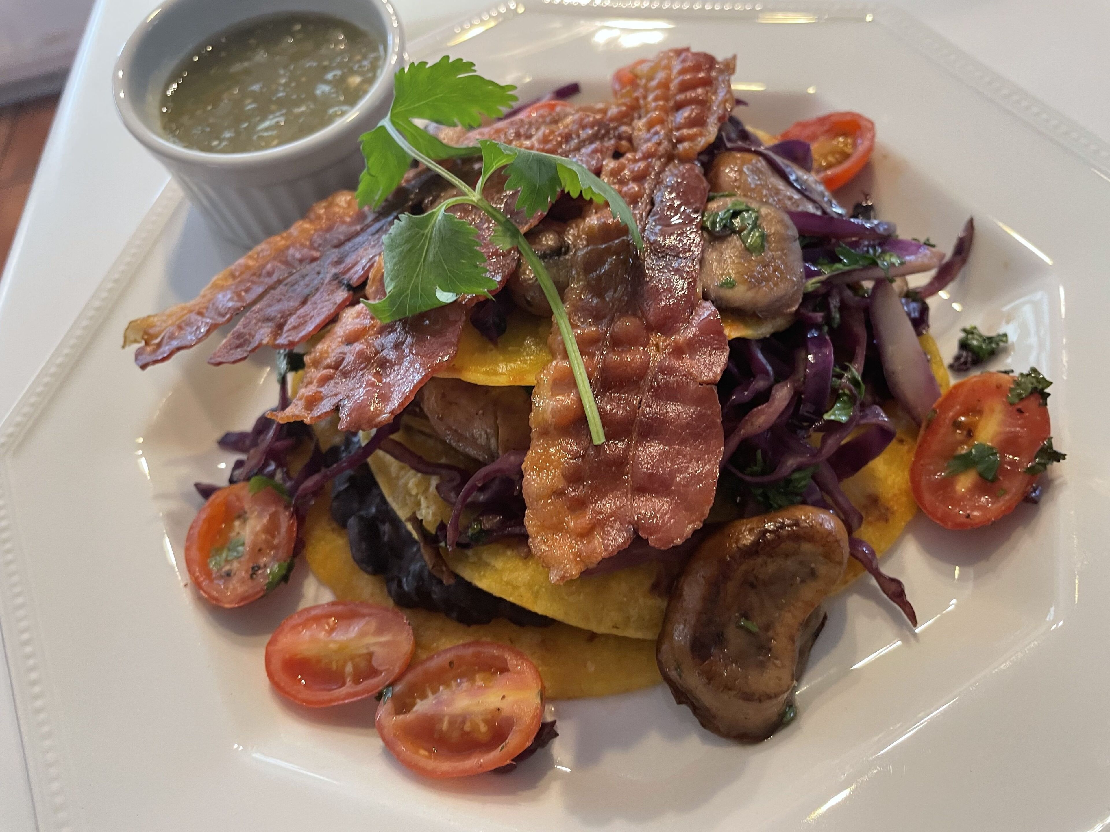 A plate of food with bacon, mushrooms, tomatoes, and greens, accompanied by a small bowl of sauce.