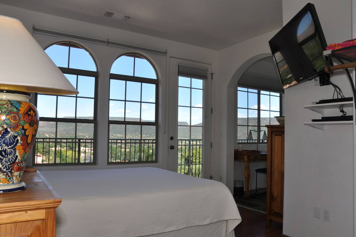 A bedroom with a large bed, a TV mounted on the wall, and a view of the outdoors through arched windows.