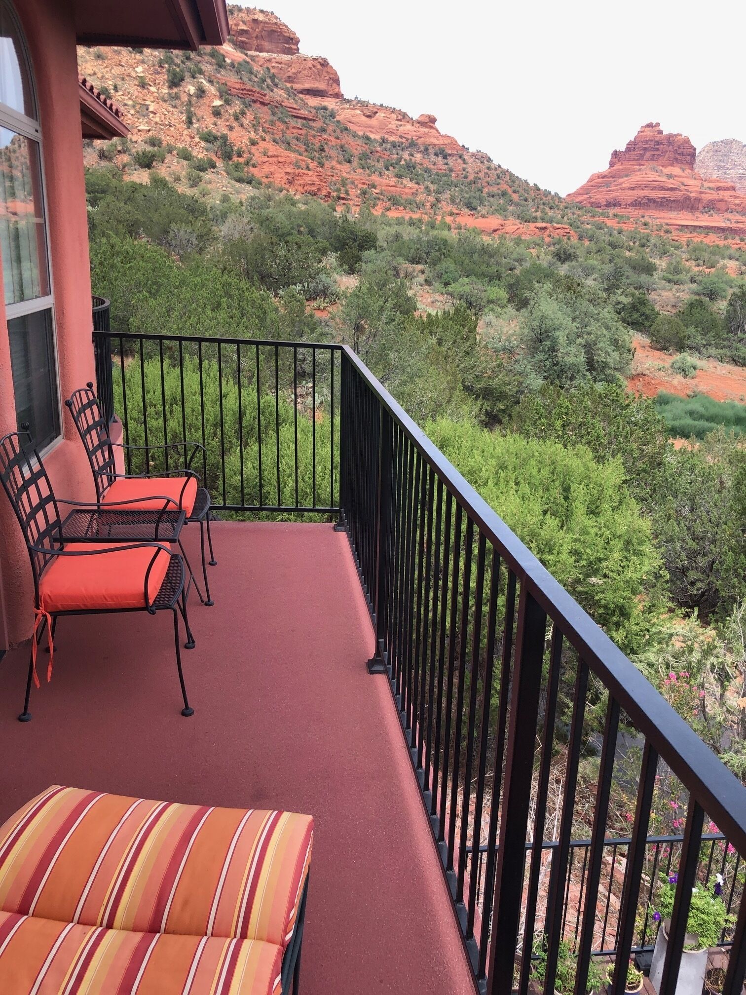 A balcony with red cushioned chairs and a striped sofa, overlooking a mountainous landscape with greenery and red rock formations.