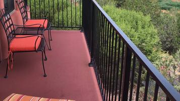 A balcony with red cushioned chairs and a striped sofa, overlooking a mountainous landscape with greenery and red rock formations.