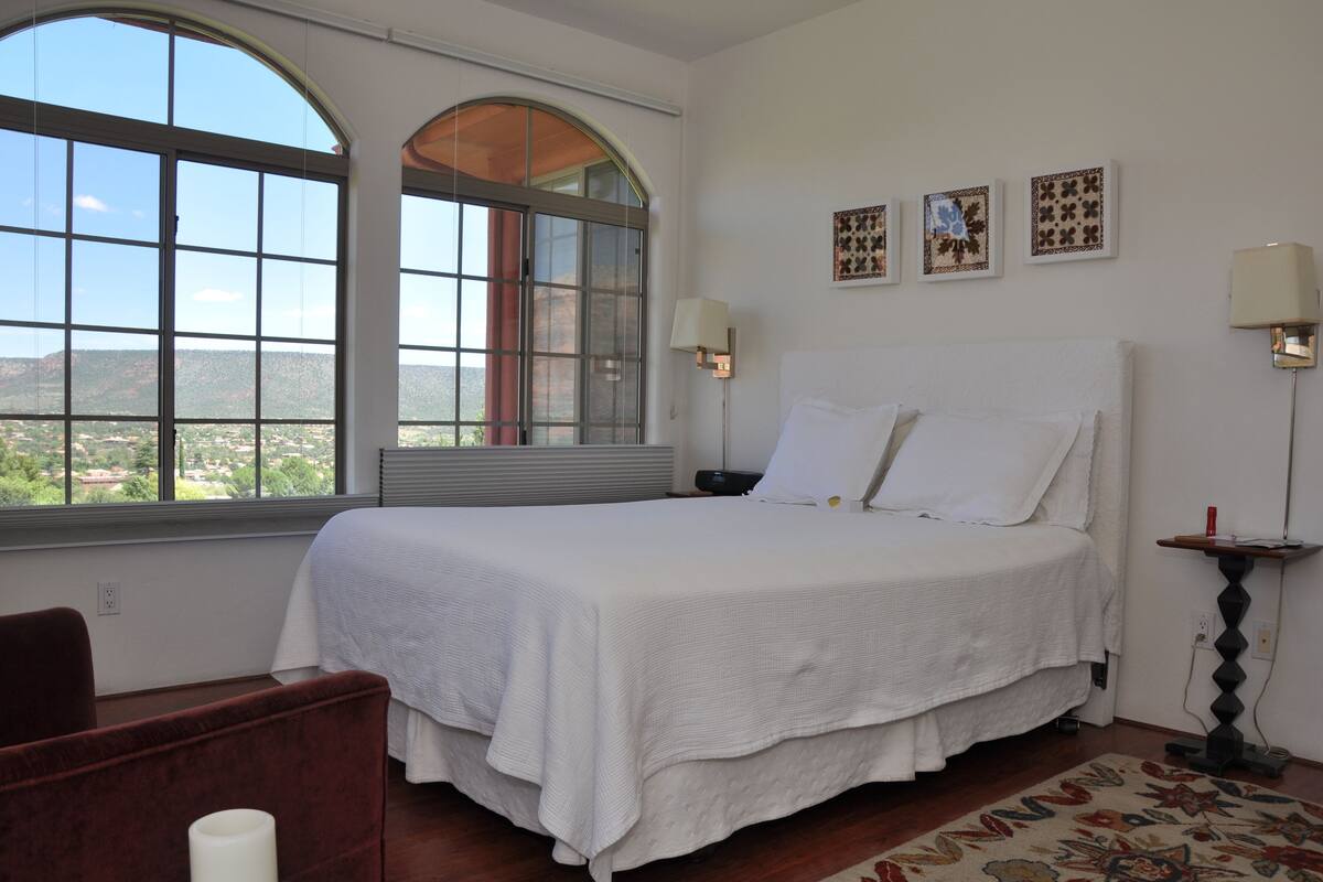 A bedroom with a large bed, a chair, a lamp, and a view of the outdoors.
