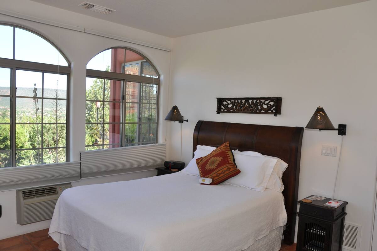 A neatly made bed with a white comforter and a decorative pillow, a nightstand with a lamp, and a large arched window offering a view of greenery.