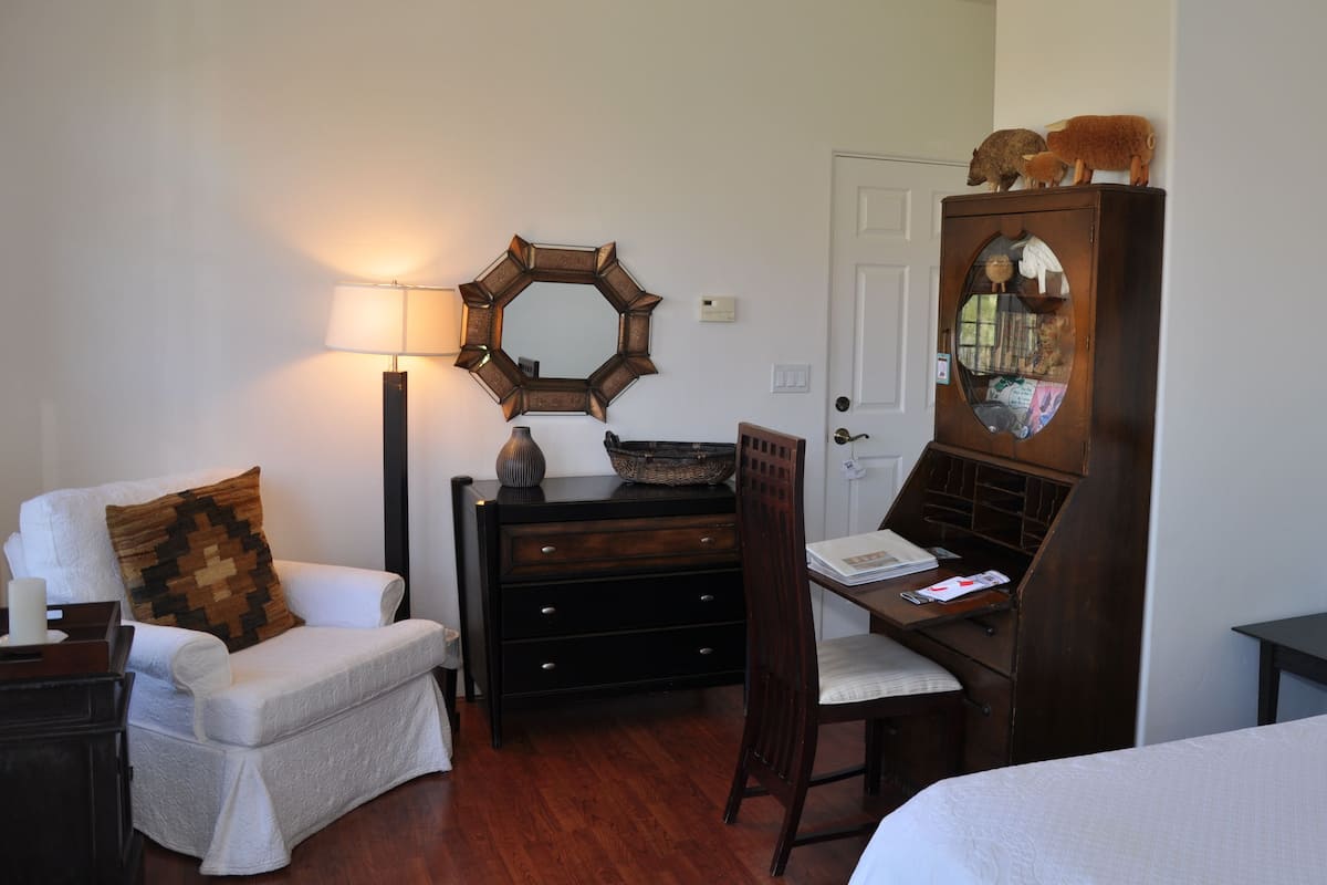 A room with a white armchair, a wooden dresser, a desk with a chair, a mirror, and a door.