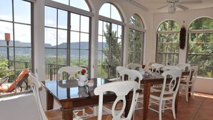 A dining area with a large table and white chairs, large windows with a view of the landscape, and a ceiling fan. - The Penrose Bed and Breakfast (Sedona)