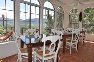 Un comedor con una mesa grande y sillas blancas, ventanas amplias con vistas al paisaje y un ventilador de techo.