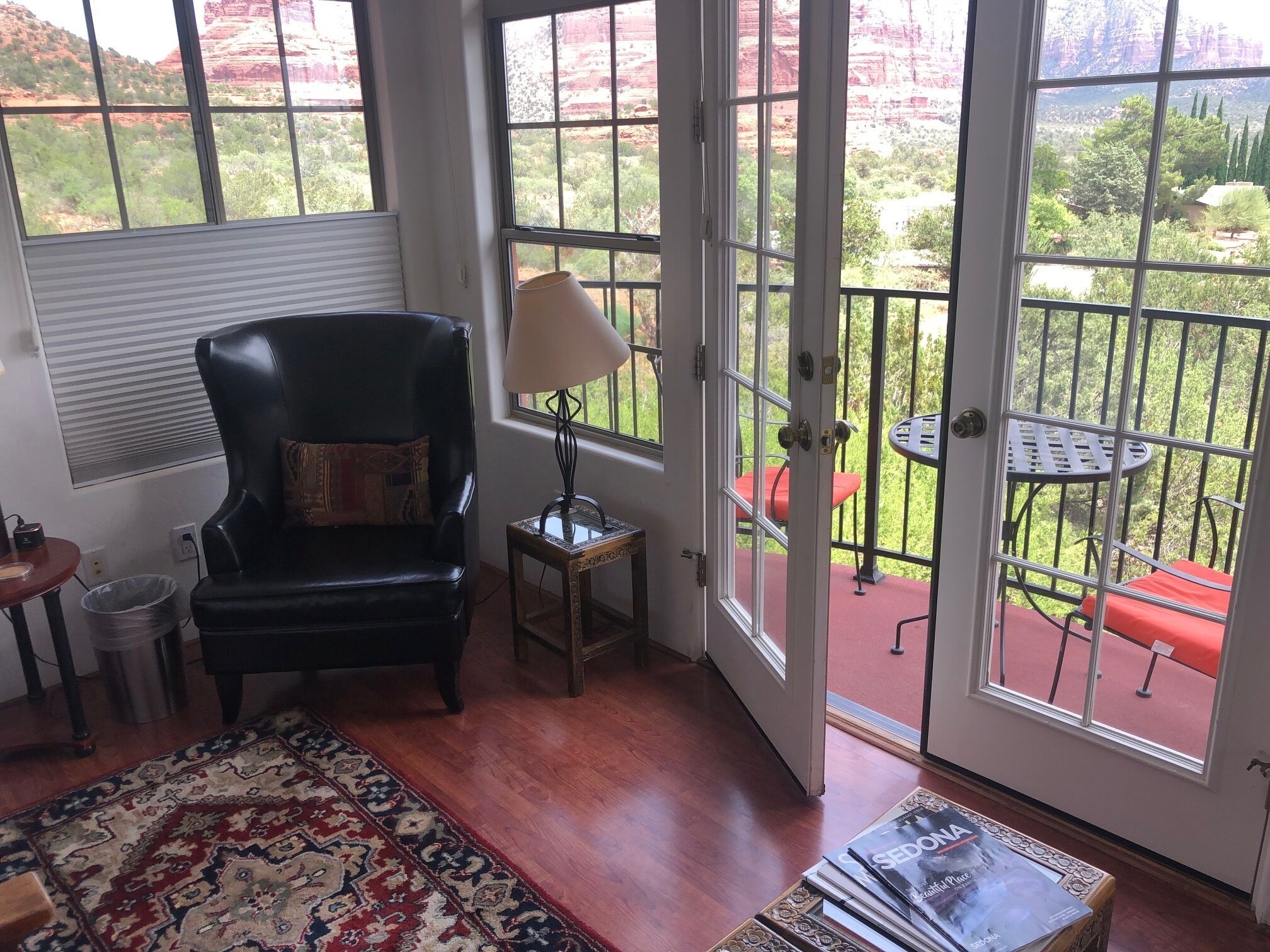 A room with a black leather chair, a patterned rug, and a table with magazines. A window reveals a balcony with a red chair and a lamp.