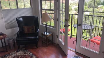 A room with a black leather chair, a patterned rug, and a table with magazines. A window reveals a balcony with a red chair and a lamp.