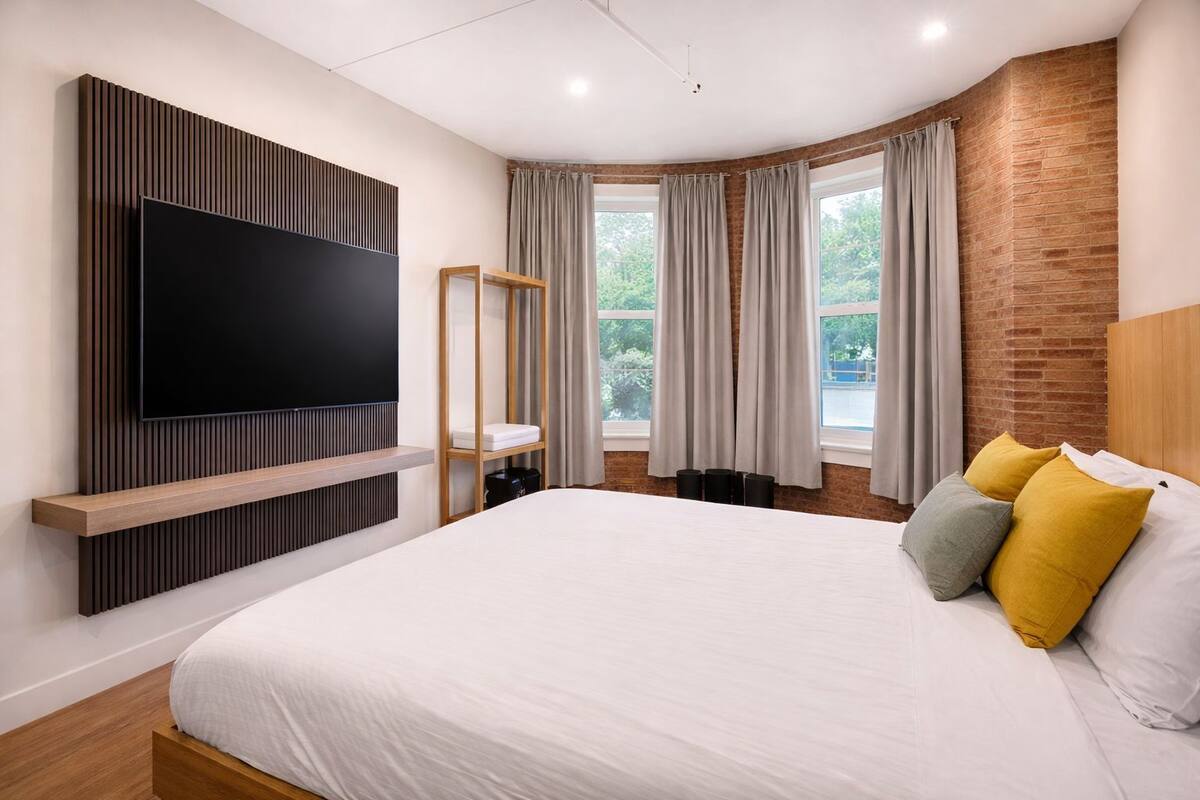 A modern hotel room with a large bed, a flat-screen TV mounted on a wooden panel, a window with curtains, and a brick wall.
