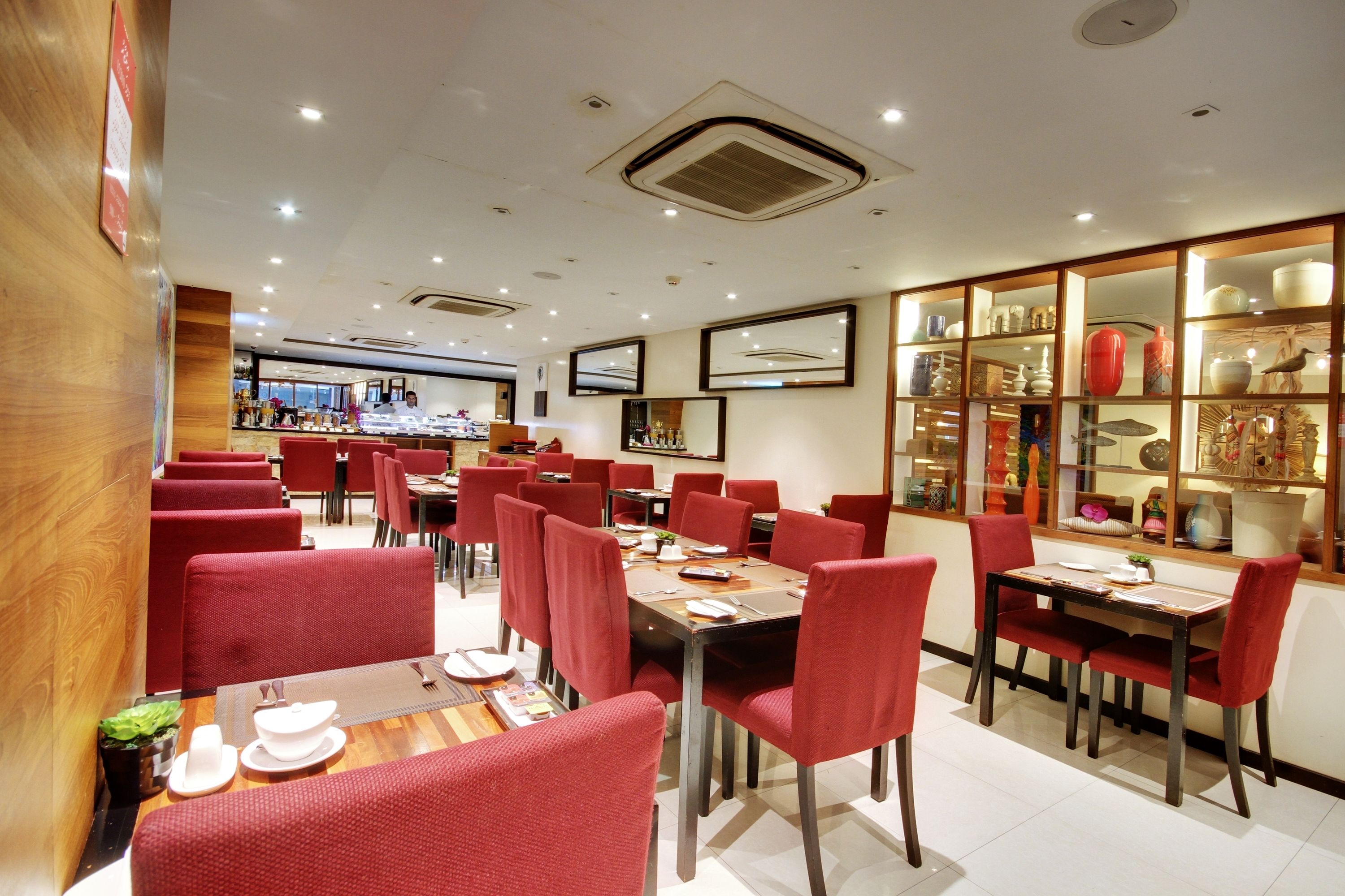 A restaurant with red upholstered chairs and tables, a display of pottery on the wall, and a counter with a variety of items.