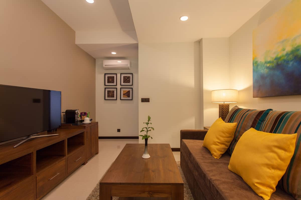 A modern living room with a flat-screen TV, wooden entertainment unit, a coffee table, a sofa with yellow cushions, and a painting on the wall.