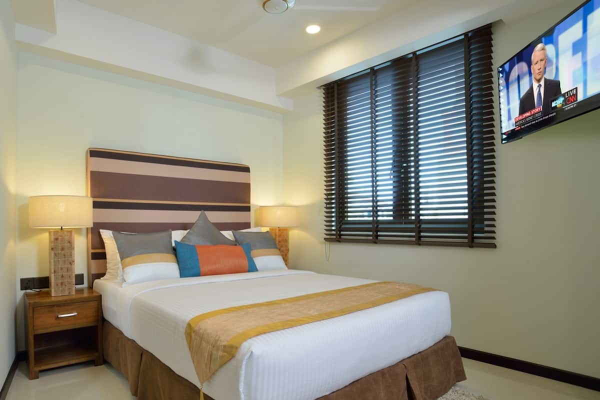 A hotel room with a large bed, two bedside tables with lamps, a flat-screen TV mounted on the wall, and a window with blinds.