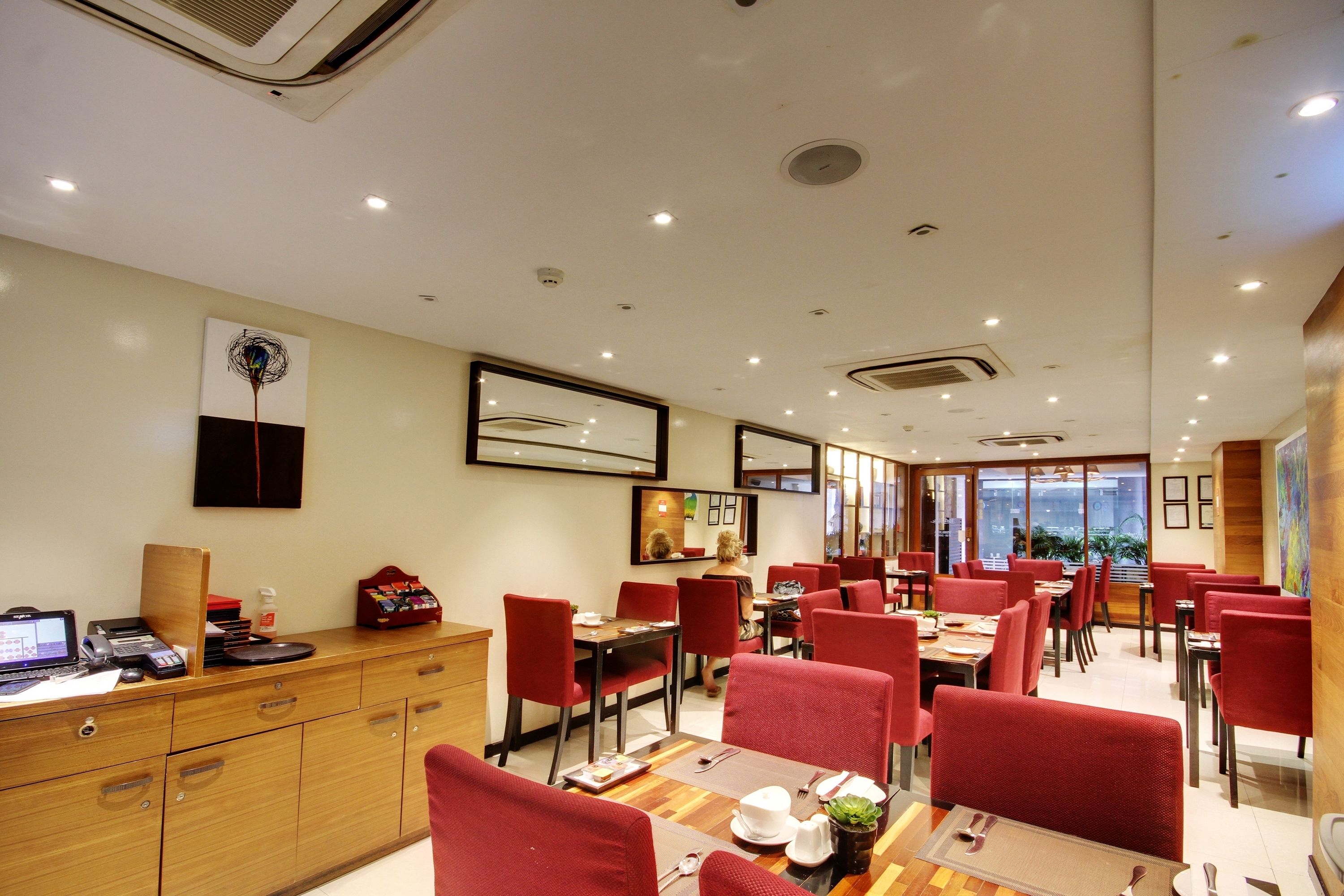 A hotel dining area with red upholstered chairs, wooden tables, and a reception desk with a computer and phone.