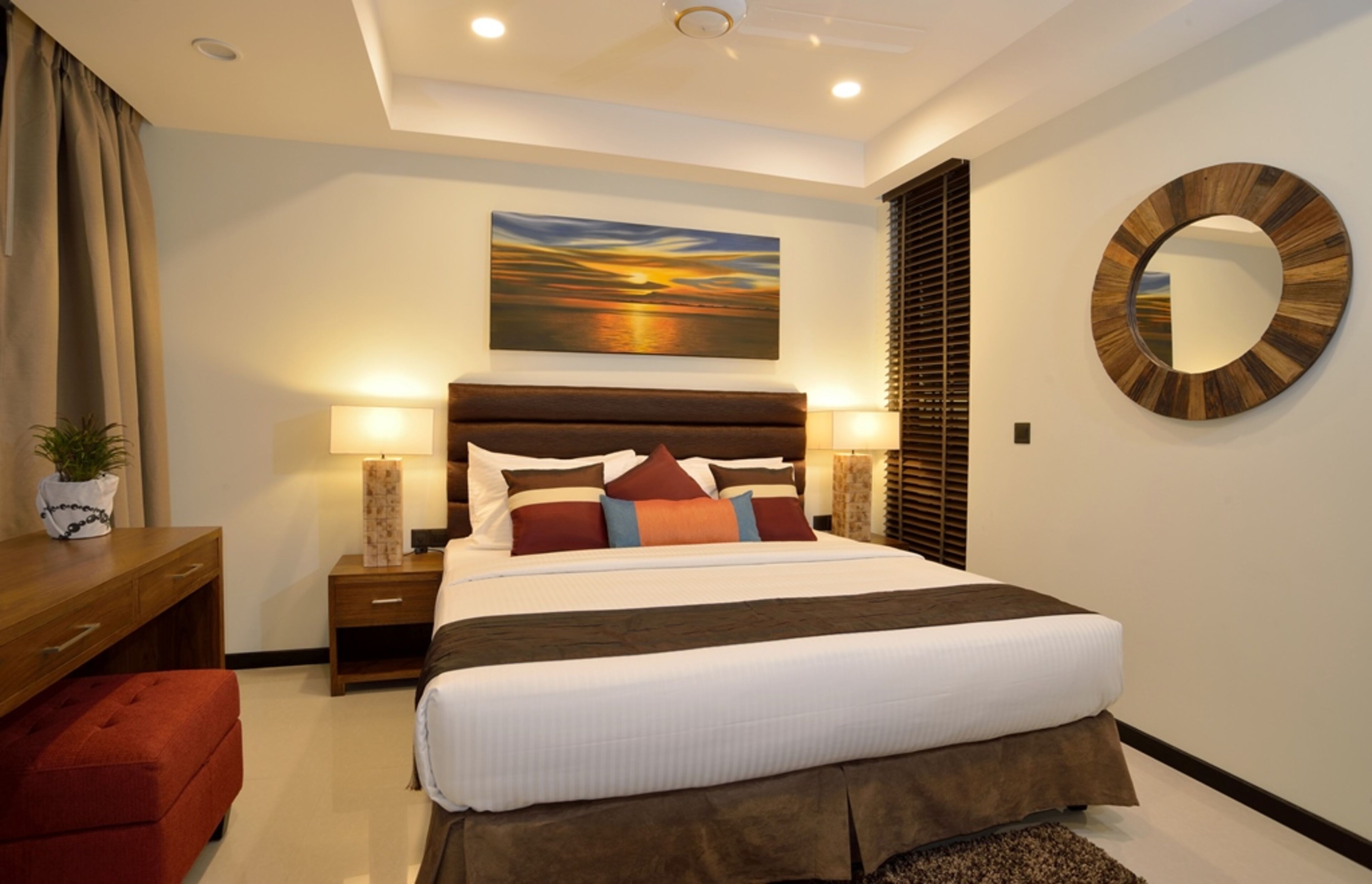A modern hotel room with a large bed, bedside lamps, a wooden headboard, a round mirror, and a painting of a sunset.