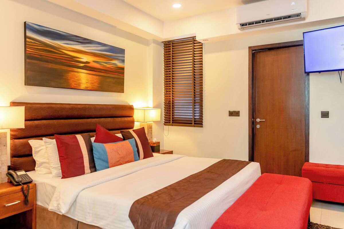 A hotel room with a large bed, a wooden headboard, a painting of a sunset, a TV mounted on the wall, and a wooden door.