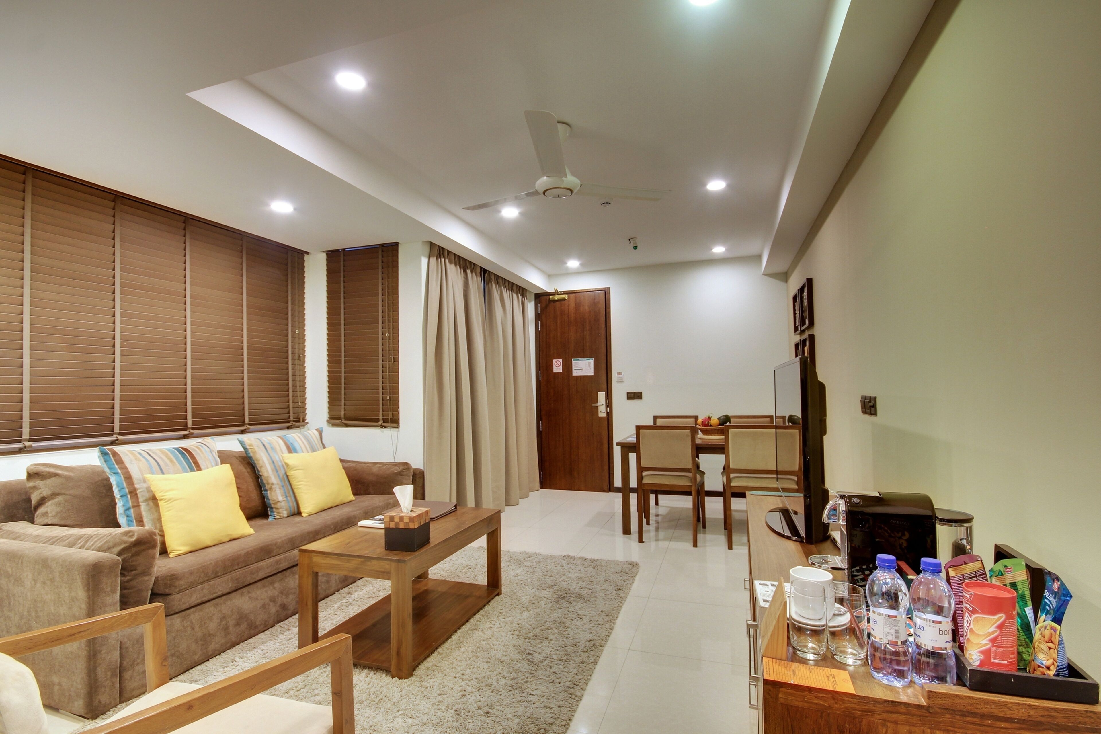 A living room with a sofa, coffee table, and a dining area with chairs and a table. There are ceiling lights and a ceiling fan.